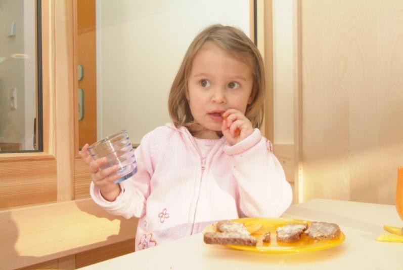 Kind in rosa Jacke mit Glas in der Hand, auf dem Tisch ein Teller mit Brot  