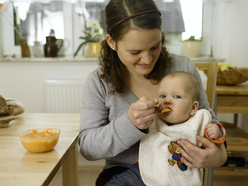 Baby sitzt auf dem Schoss, Mutter füttert es mit Löffel 